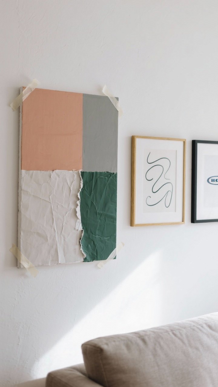 Closeup detail shot of a small gallery wall in a bright living room, showing a color-block canvas with taped geometric shapes in three tones (clay, greige, and forest green), a textured paper collage with torn edges layered under a white mat, and an abstract squiggle print in a thrifted gold frame beside an IKEA black frame; art hung at eye level on a clean white wall, soft natural daylight, subtle wall texture, crisp edges where painter’s tape was removed, cohesive palette limited to three tones